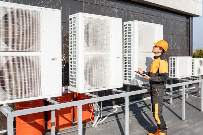 A man in black and yellow construction gear holding a clipboard and pen taking notes on the commercial HVAC equipment to see what kind of HVAC maintenacne the business will need